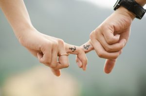 zwei Menschenhalten sich an den Zeigefingern, auf denen jeweils ein Anker tättowiert ist