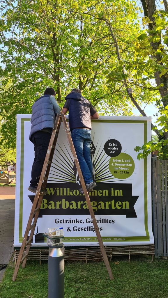 Zwei Personen stehen auf einer Leiter und hängen ein Plakat aus.