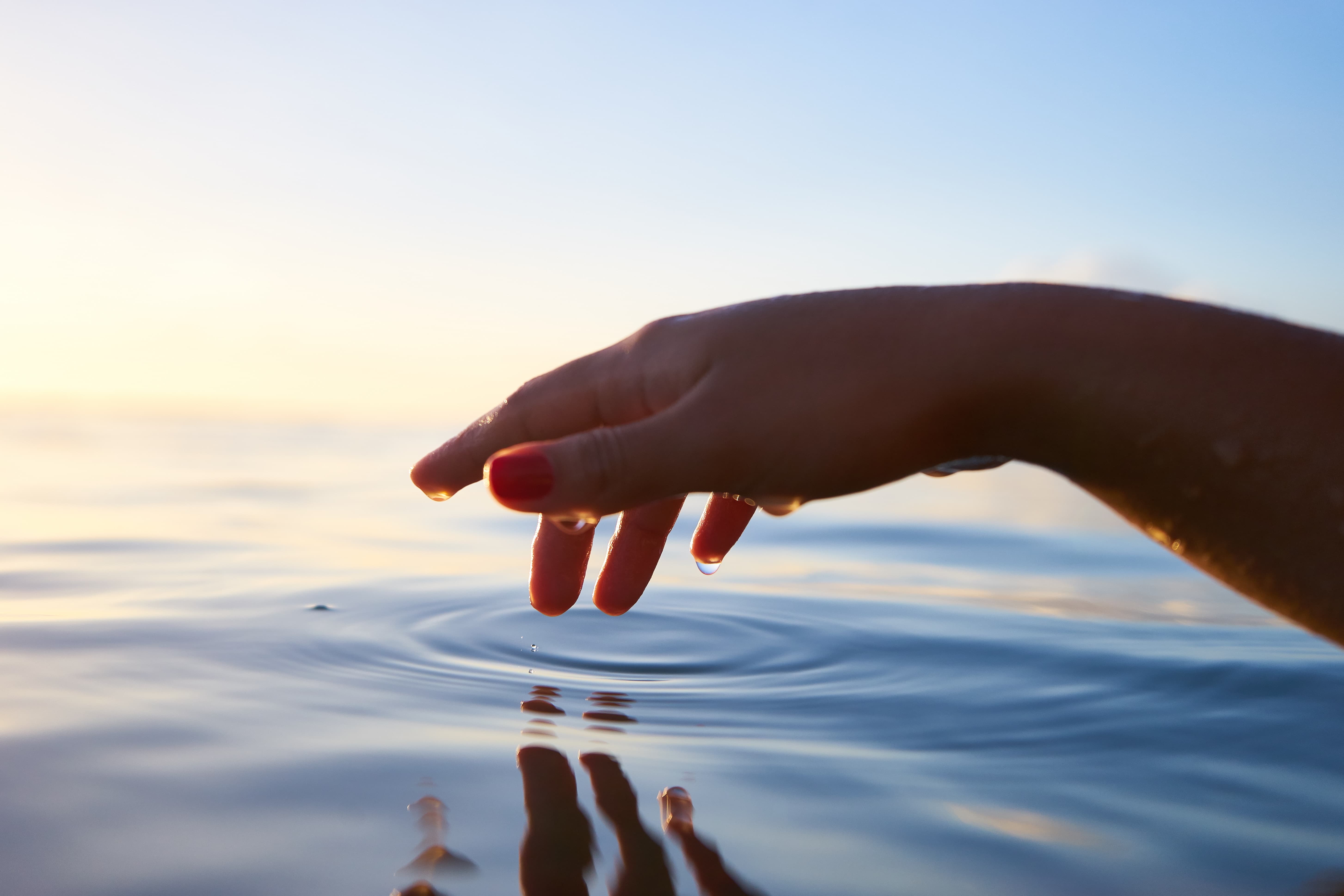 Eine Hand gleitet über eine glatte Wasseroberfläche und hat diese scheinbar kurz zuvor berührt. Ruhe und Heilung schwingen mit.