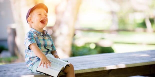 Junge sitzt mit einem Buch auf einem Steg und lacht