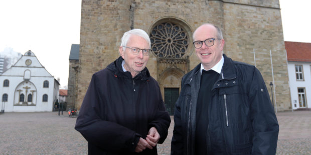 Zwei dunkel gekleidete Männer stehen vor einer Kirche.