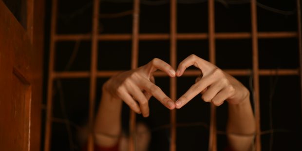 Eine Person hinter Gitterstäben zeigt mit den Fingern ein Herz.